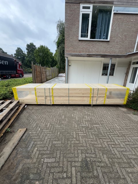 Large wooden beam wrapped in yellow straps on a driveway outside a brick house