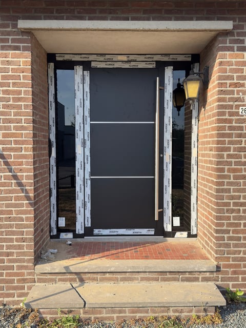 Black front door under construction with white-painted frame in red brick doorway, yellow light fixture visible on right side